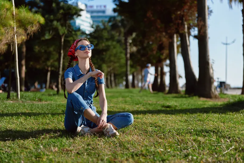 Lifestyle woman in modern boho-western denim street style sits on grass under trees wearing sunglasses and red bandana, enjoying sunny day with film color tones.
