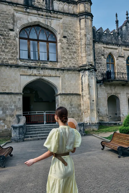 Castle courtyard scene with a woman in a pale yellow dress seen from behind, exploring ancient stone arches in a timeless travel moment.