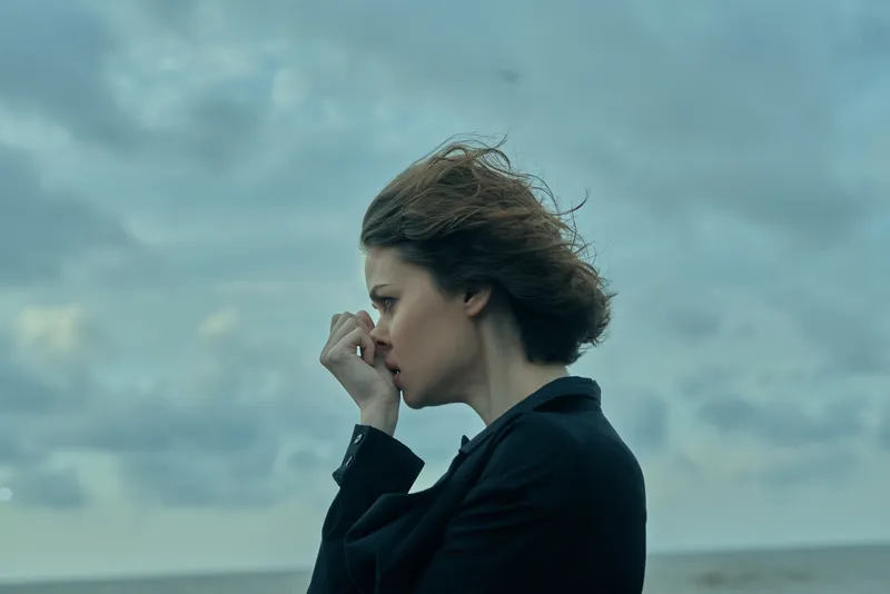 Woman in a dark coat stands by the sea, wind tousling hair under a cloudy moody sky, capturing a contemplative outdoor moment with dramatic coastal weather.