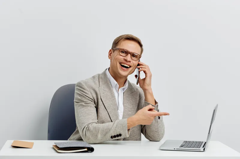 Smiling mature man with glasses talking on mobile phone while sitting at a desk in an office environment, using a laptop and taking notes, indoor shot with neutral background. Business lifestyle conce