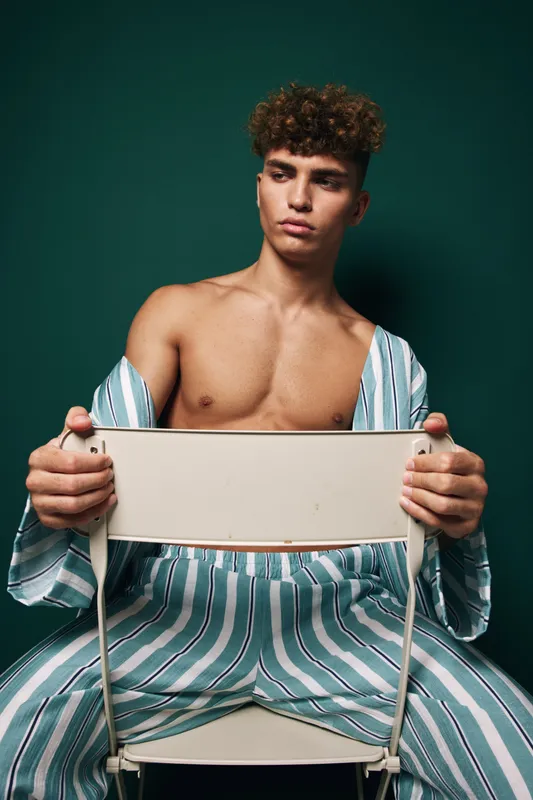 Young man with curly hair wearing striped pajamas sitting on chair backwards with serious expression on dark green background in studio.