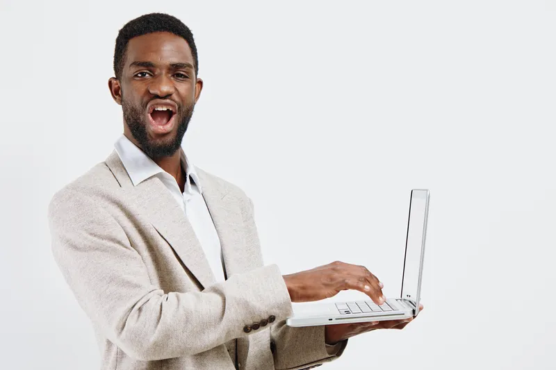 Smiling man with dark skin and short curly hair holding a laptop, wearing a beige blazer and white shirt, presenting cheerfully against a plain white background, studio portrait, people lifestyle conc