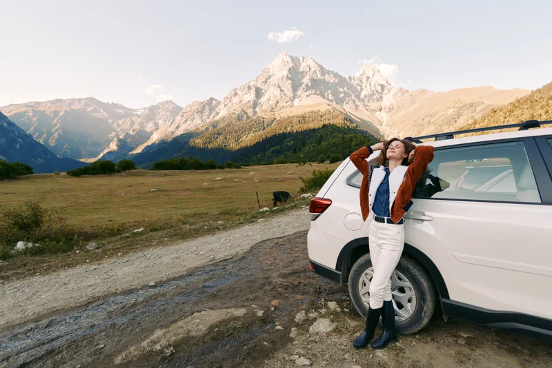 Woman, car, mountains, road, landscape, travel, nature, relaxation. Young woman leaning against a white SUV on a gravel roadside with towering peaks and sweeping valley views outdoors.
