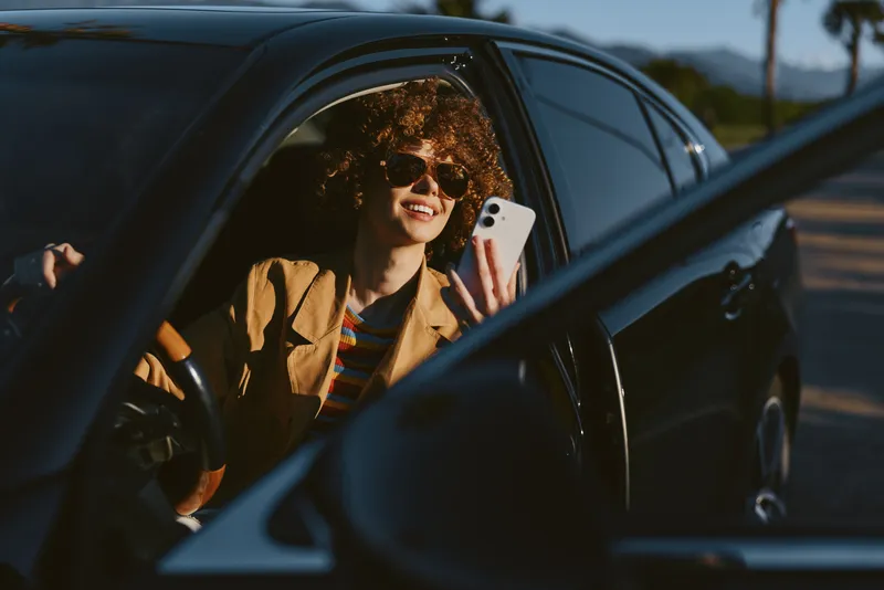 Cheerful woman inside a car, smiling and relaxed, sunlit road scene with warm retro film color, candid moment, casual outfit, natural lighting, vibrant lifestyle, carefree attitude