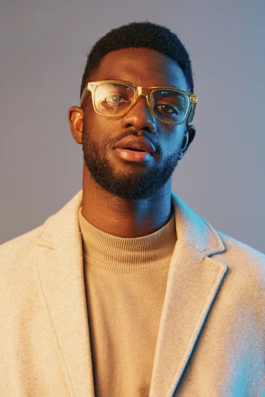 Stylish young man with glasses in a beige coat, showcasing confidence against a gradient background, ideal for modern fashion concepts and lifestyle branding