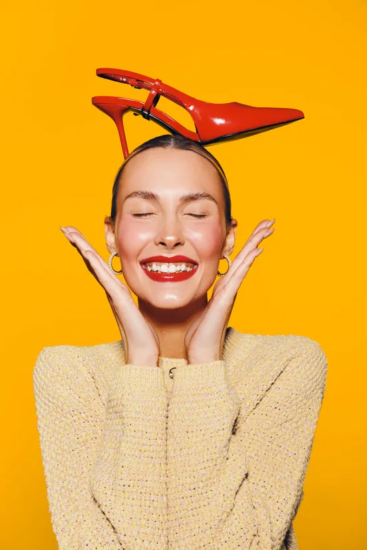 Happy woman with red lipstick and earrings smiles with closed eyes, holding red high heel shoes on her head against bright yellow background. Concept of joy and fashion.