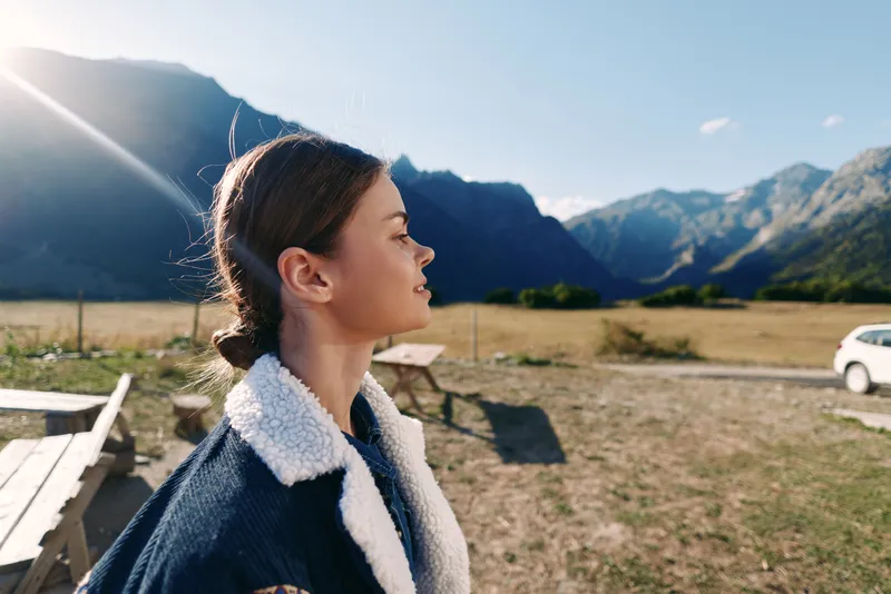 girl child profile mountains nature sunlight outdoors portrait of a young girl in a shearling jacket standing in a meadow by a road, serene expression, sunbeam and scenic valley background