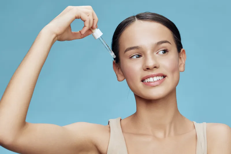 Young woman applying facial serum with a dropper in a light blue background, promoting skincare and healthy beauty routines