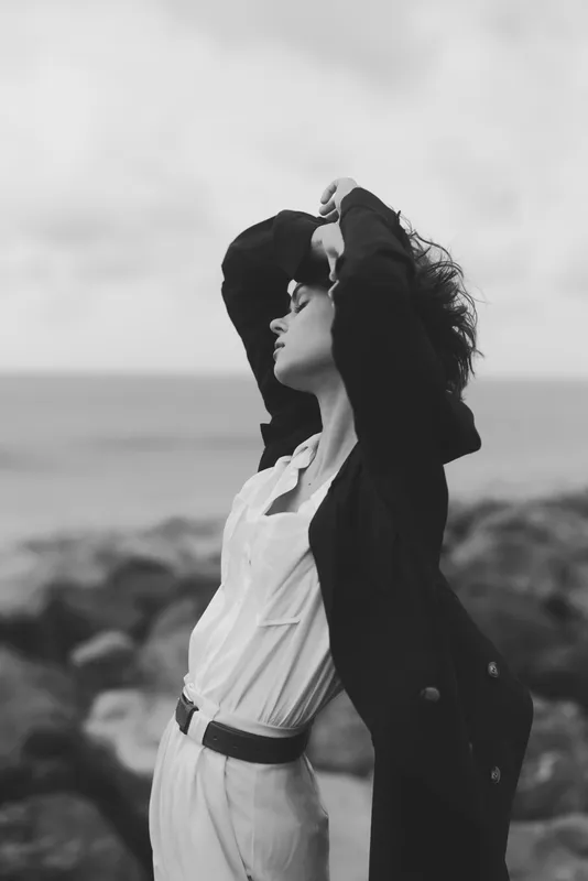 Fashionable woman posing by the sea in a white shirt and black coat, wind swept hair, monochrome photography, elegant lifestyle, outdoor styling with coastal atmosphere