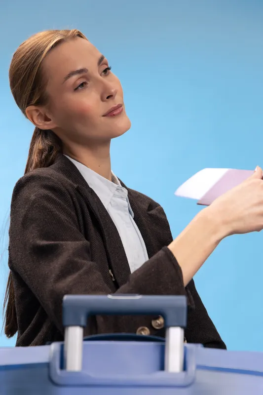 Young woman in black blazer holds passport and boarding pass near suitcase handle at airport. Concept of travel, business trip, and female traveler preparing for flight.