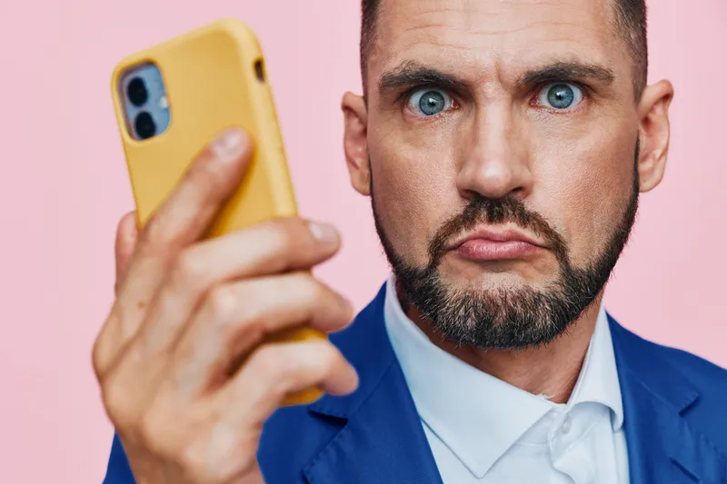 A professional man in a blue suit studies his smartphone with a focused, puzzled expression against a bright pink background, creating a clean, isolated color aesthetic ideal for modern business
