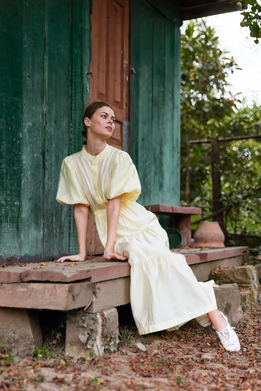 woman outdoor fashion seated on steps in a garden, wearing a pale dress, serene pose beside a green wooden wall, natural daylight and calm atmosphere