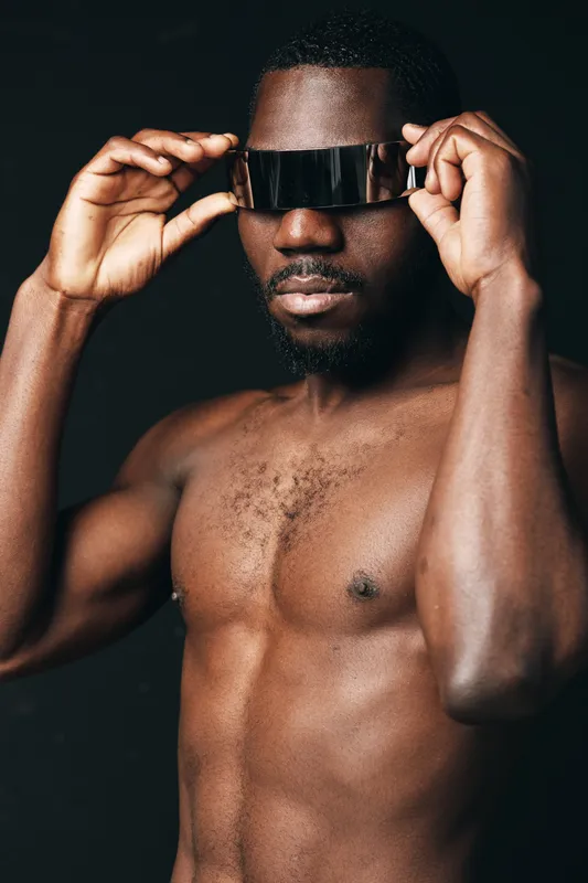African man shirtless wearing futuristic visor glasses in creative conceptual visual style with dark background. Portrait of confident black male model with modern tech accessory.