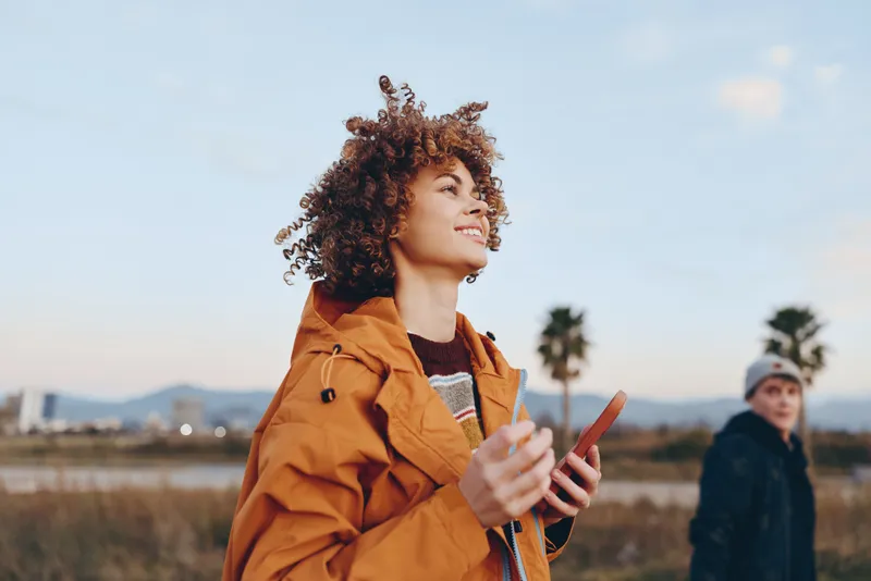 woman, lifestyle, outdoors, curly, hair, orange, coat, sunset, field, smiles while holding phone and sunglasses, warm evening light, countryside meadow, blurred friend in background, candid travel