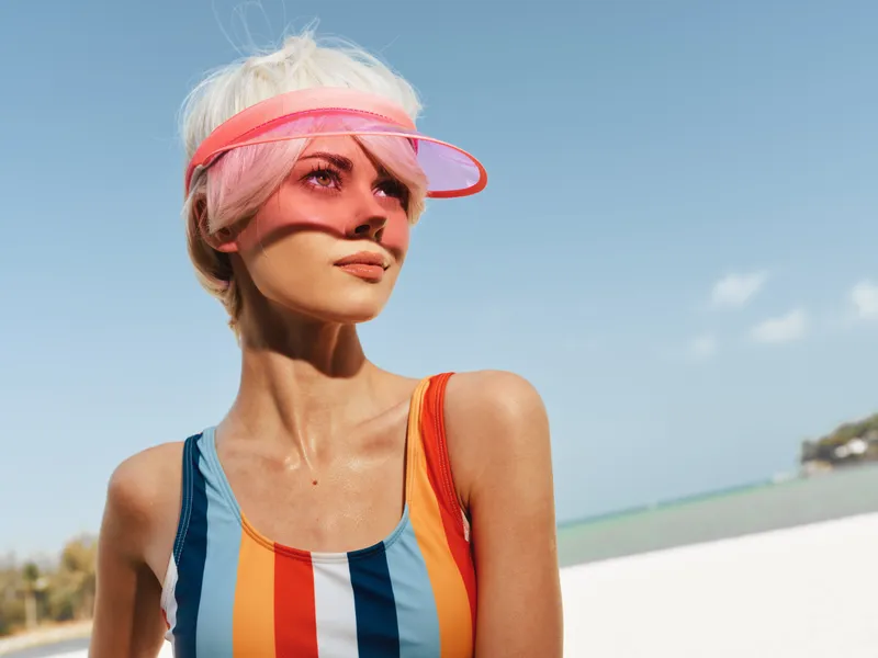 Portrait of a stylish woman wearing a colorful striped swimsuit and a translucent pink visor, standing on a sunny beach with clear blue sky and calm sea.