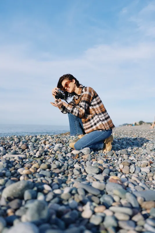 Photographer kneels on a pebble beach, aiming a camera toward the sea, capturing an outdoor portrait and seascape under a clear blue sky.