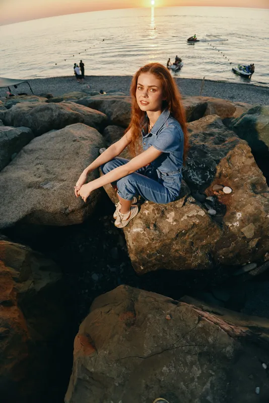 Modern boho-western denim street style woman with red hair sitting on rocks by the sea during sunset, captured with warm film color tones for a casual lifestyle vibe.