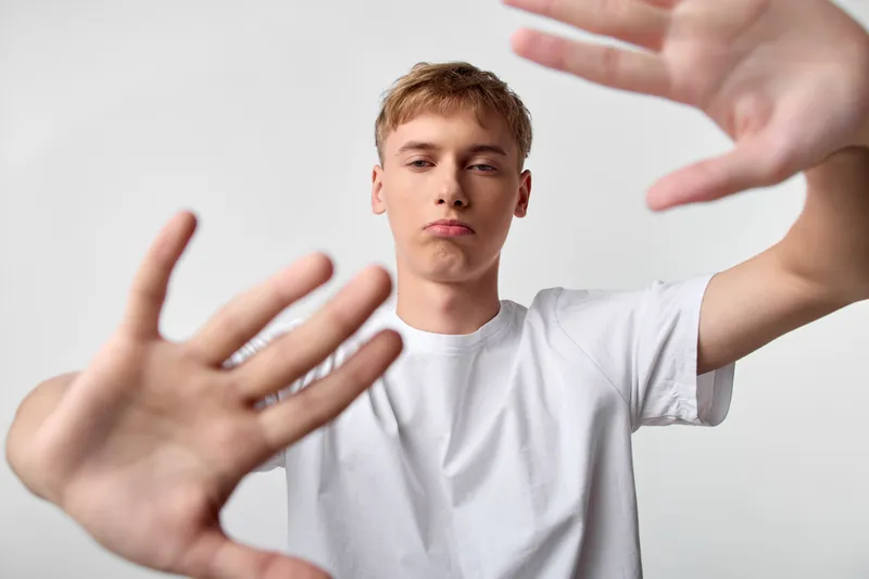 Hands reach toward camera in front of a focused youth wearing a white tee, creating distance, barrier and tension in a minimal studio setting