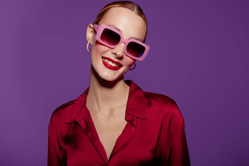 Smiling woman in pink sunglasses and red blouse poses against purple background. Fashion portrait with bright colors and confident mood. Concept of style and beauty.