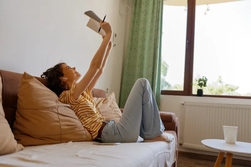 Cheerful woman indoors, lounging on a sofa, reading a book, relaxed posture, soft sunlight, cozy home atmosphere, natural colors, casual lifestyle, warm vibe
