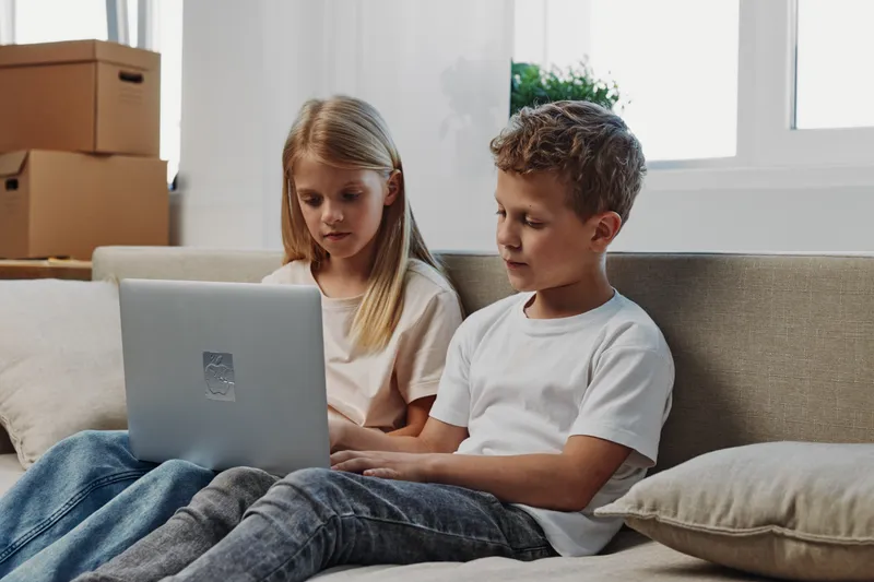 Two focused children, girl and boy, using laptop on sofa in living room with cardboard boxes. Concept of learning, technology, and home environment.