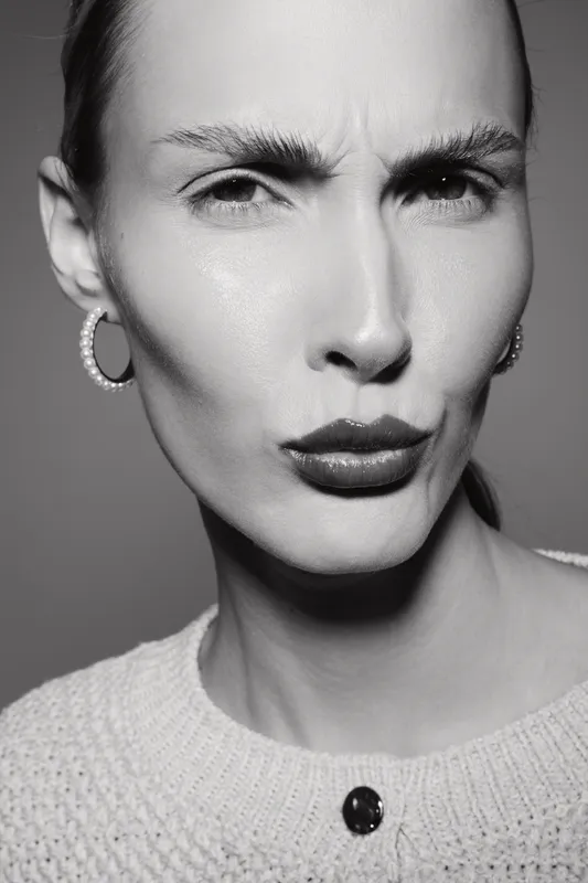 Close-up of a young white woman with pale skin making a skeptical facial expression. She wears a knitted sweater and pearl earrings in a black and white studio portrait.