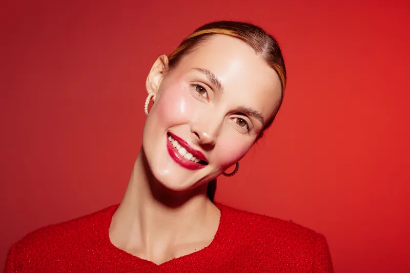 Smiling woman with shiny skin and red lipstick poses against red background wearing red sweater. Concept of beauty, makeup, skincare, and fashion with vibrant color contrast and studio lighting.