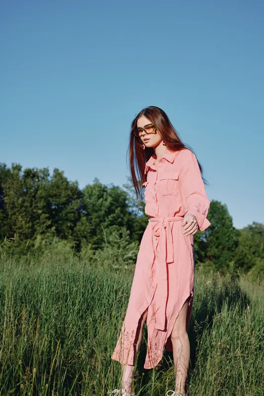 Stylish young woman in a flowing pink dress walking through a green field on a sunny day, radiating confidence and joy while enjoying nature