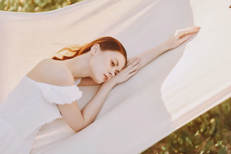 Dreamy young woman in a white dress resting on a sunlit hammock, showcasing tranquility and serenity amidst a lush, vibrant landscape