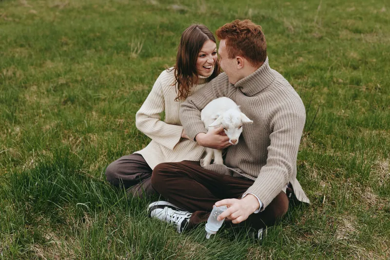 Cheerful couple outdoors, smiling together while holding a lamb, warm sweaters, sunlit moment, gentle breeze, natural colors, grass and meadow background, relaxed pose, affectionate mood, lifestyle