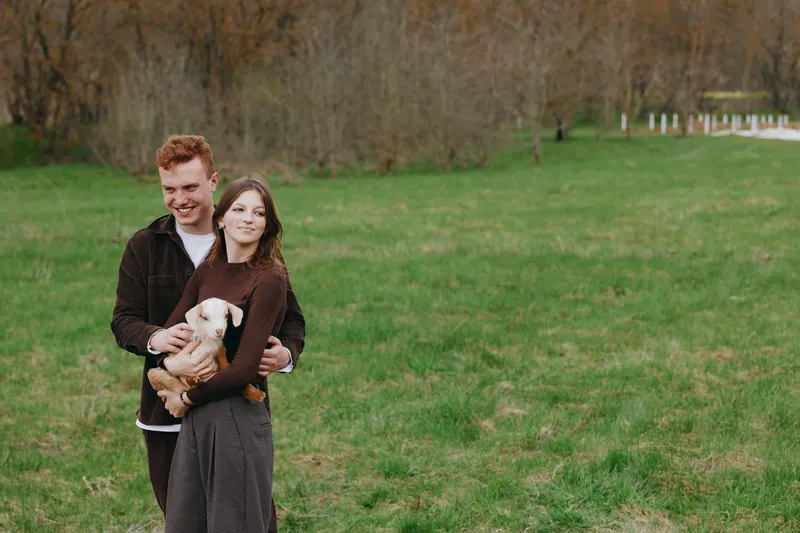 Smiling couple outdoors in a grassy park embracing a small dog, sunlight casting warm tones. Honest, cozy lifestyle shot showcasing companionship, affection, and everyday happiness in natural