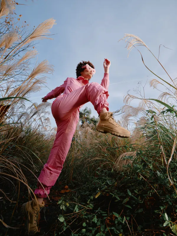 Outdoor action jump, a person in a pink jumpsuit leaps above tall grasses in a natural landscape, capturing bold movement and energy with a sense of freedom under a bright sky