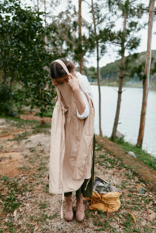 A young woman in a serene outdoor setting holds a beige dress by the lake, expressing a contemplative mood surrounded by nature and greenery