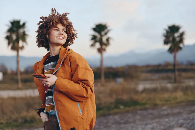 Woman lifestyle outdoors scene, curly hair, orange jacket, scarf, walking along rural path at golden autumn sunset, soft relaxed expression, natural background with palm trees and open sky