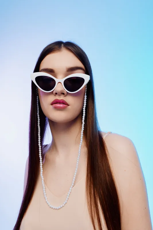 Young woman with long hair wearing white fashion sunglasses and pearl chain poses against blue background. Concept of beauty, fashion, style, and accessories for modern trends.