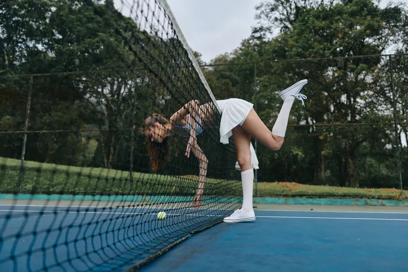 Young female tennis player in sporty attire stretching over the net on a sunny day, portraying determination and energy in a lush green outdoor setting