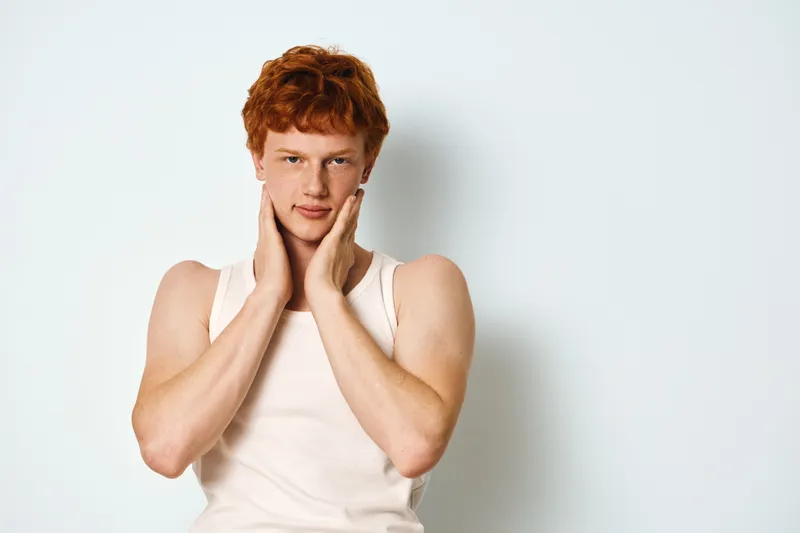 Young man with red hair in white tank top posing with hands on neck looking at camera on white background. Studio portrait with casual style and confident expression.