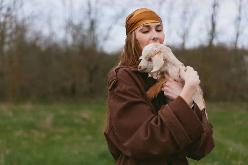 A woman in a brown coat and orange headband cuddles a friendly goat kid in a grassy field, capturing rural lifestyle, bonding, and animal companionship under soft light, warmth together