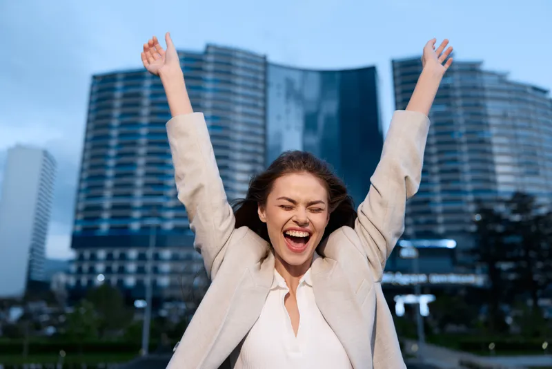 Young cheerful woman raising hands celebrating success outdoors near modern office buildings in business district urban environment