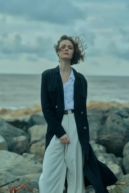 Woman on a rocky beach wearing a long dark blazer over a light shirt and wide trousers, wind tousling hair, moody seaside atmosphere, fashion shoot for outdoor apparel.