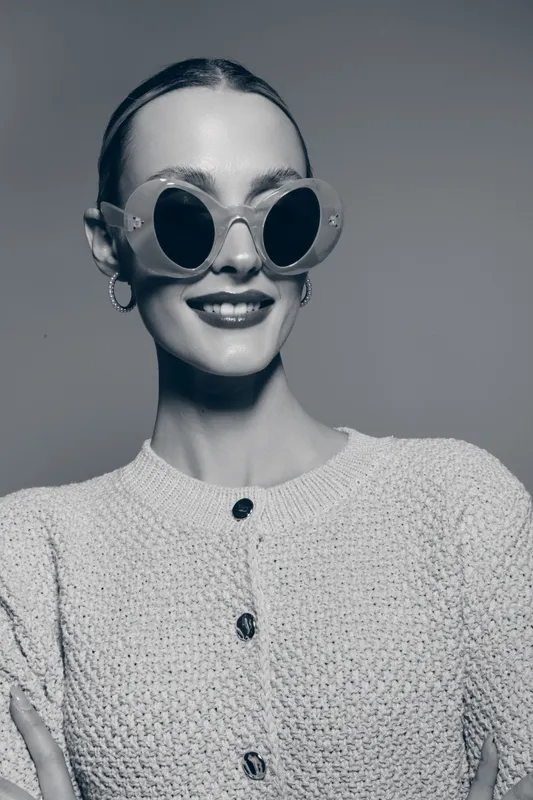 Young white woman with pale skin wearing round sunglasses and knitted sweater smiles confidently. Studio portrait with minimalistic background and modern fashion style concept.