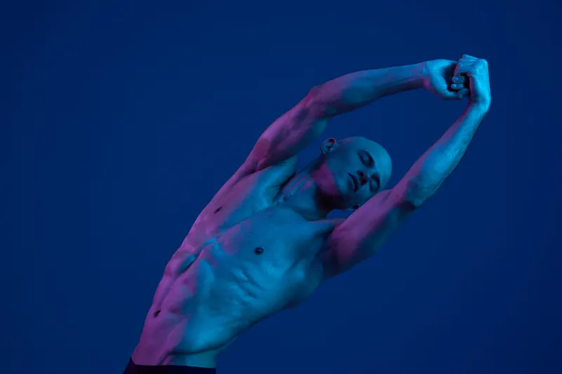 Strong man stretching under colorful light, showcasing well-defined muscles and an expressive pose. The vivid colors create a dynamic and energetic atmosphere.