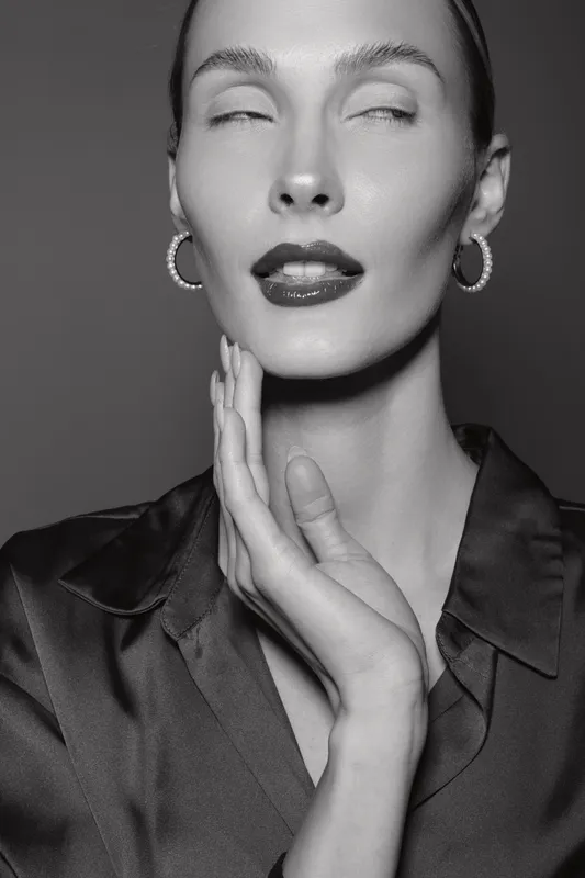 Elegant Caucasian woman with pale skin poses in black satin blouse with closed eyes and hand near face. Studio portrait with soft light and glamorous mood.