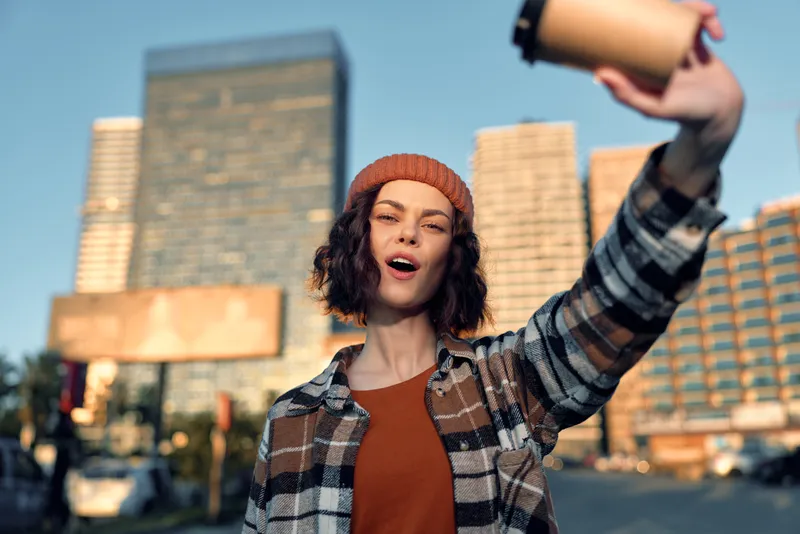 Woman portrait holding coffee selfie in urban street, candid smile and beanie at golden hour. Emotional storytelling, authenticity and mindful living in a modern candid lifestyle scene.
