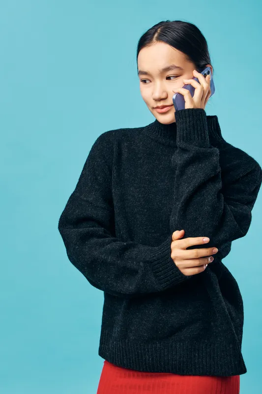 Woman talking on phone with thoughtful expression wearing black sweater and red skirt standing against blue background in studio. Communication lifestyle and casual fashion portrait.