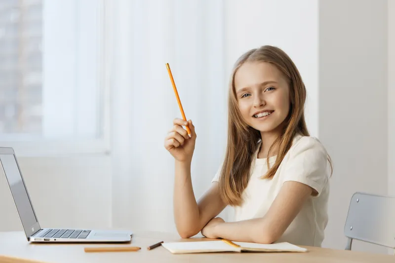 Smiling young girl with a pencil at home study desk, dressed casually in white, exuding confidence and happiness in a bright, airy environment