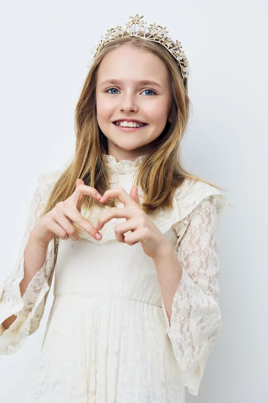 A young girl in a delicate lace dress and crown, smiling joyfully, making a heart shape with her hands against a soft, neutral background