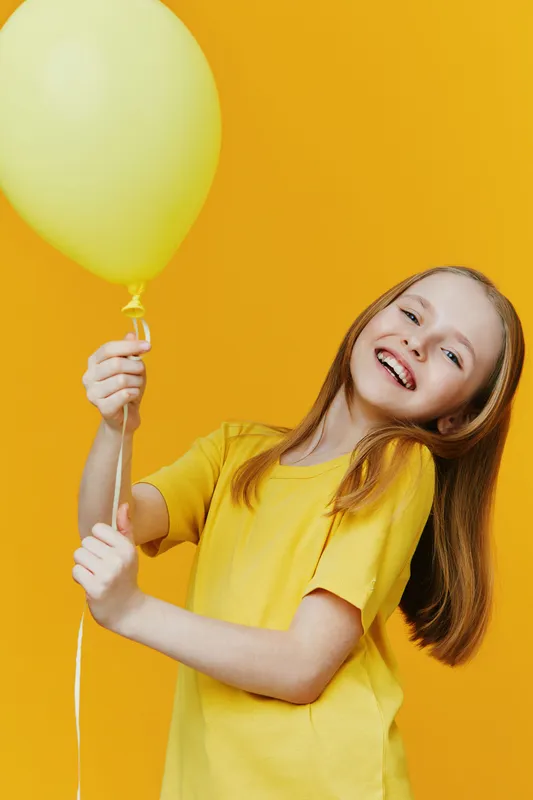 Happy girl with long hair holding a yellow balloon, wearing a yellow shirt against a vibrant yellow background, conveying joy and playfulness in a cheerful concept