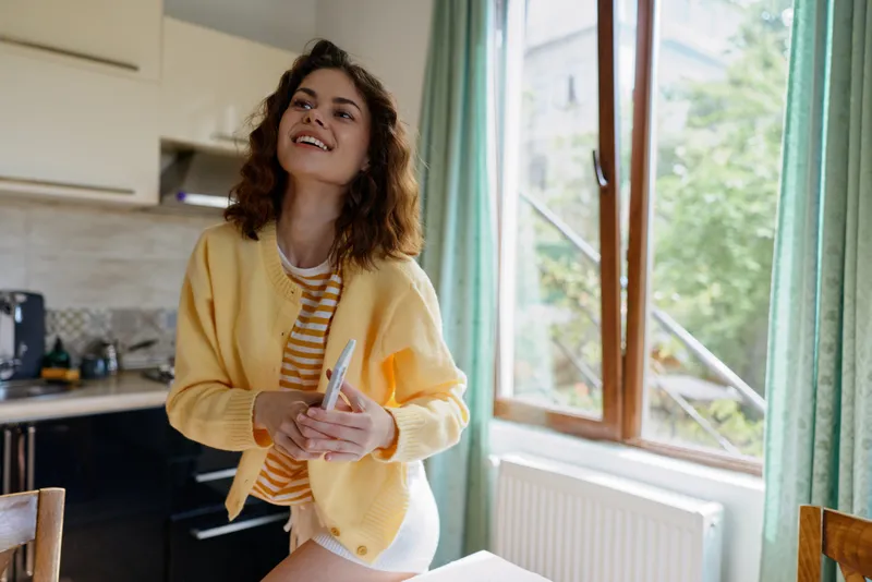 Cheerful woman stands in a bright kitchen wearing a yellow cardigan, holding a utensil, enjoying a casual morning at home, smiling at the camera under warm natural light