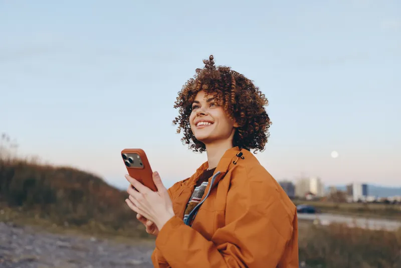 Woman lifestyle portrait outdoors at sunset, smiling with curly hair, holding a smartphone and looking at the screen, casual orange jacket, calm riverside background, travel mood.
